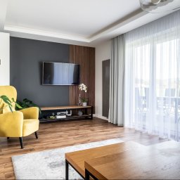 Bonus Nieruchomości - Jasne wnętrze salonu z żółtym fotelem, telewizorem na ścianie z drewnianymi panelami i dużym oknem z widokiem na zewnątrz, podkreślone naturalnym światłem.
