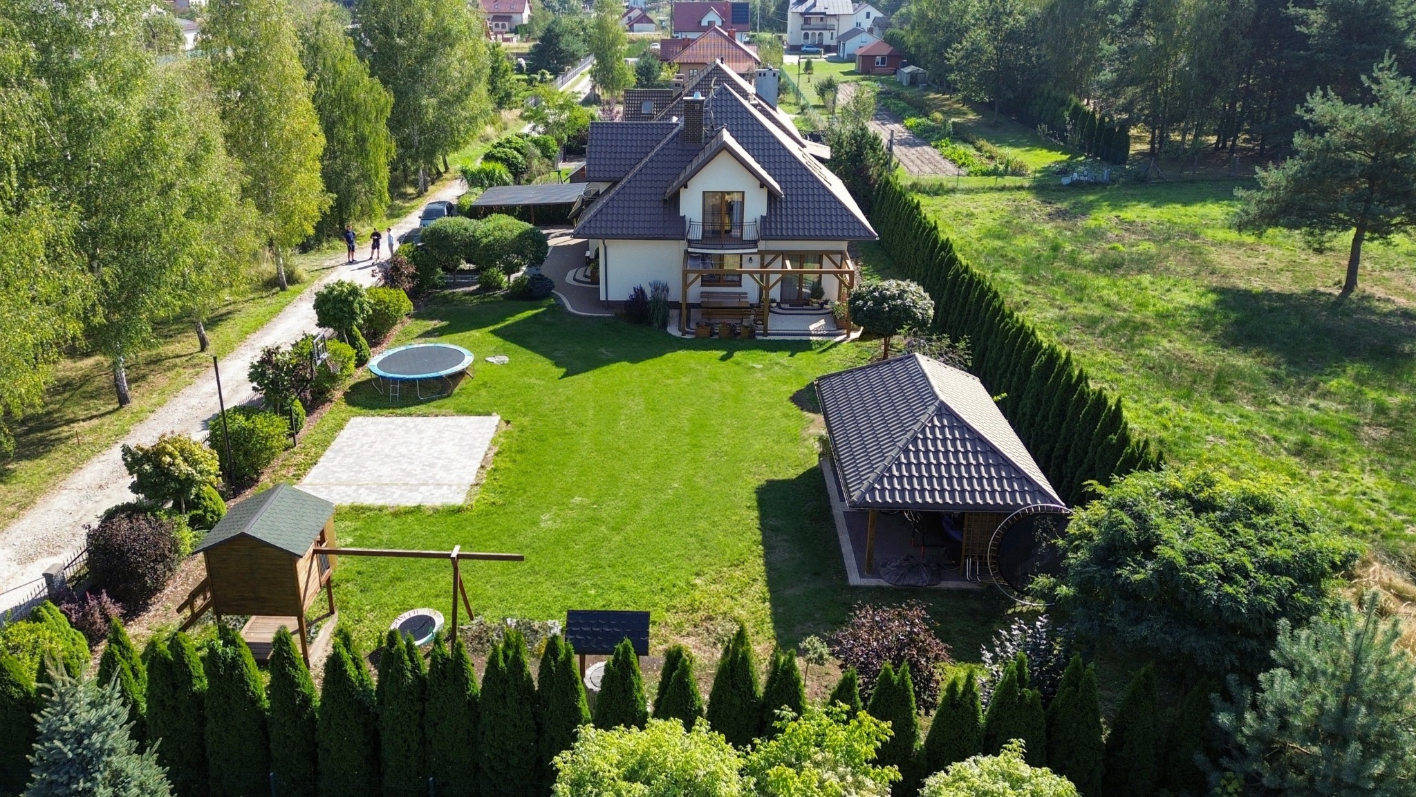 Widok z lotu ptaka na dom z ogrodem, gdzie widać trampolinę, plac zabaw, altankę i zadbany trawnik. Widoczne inne domy w oddali, słoneczny dzień.