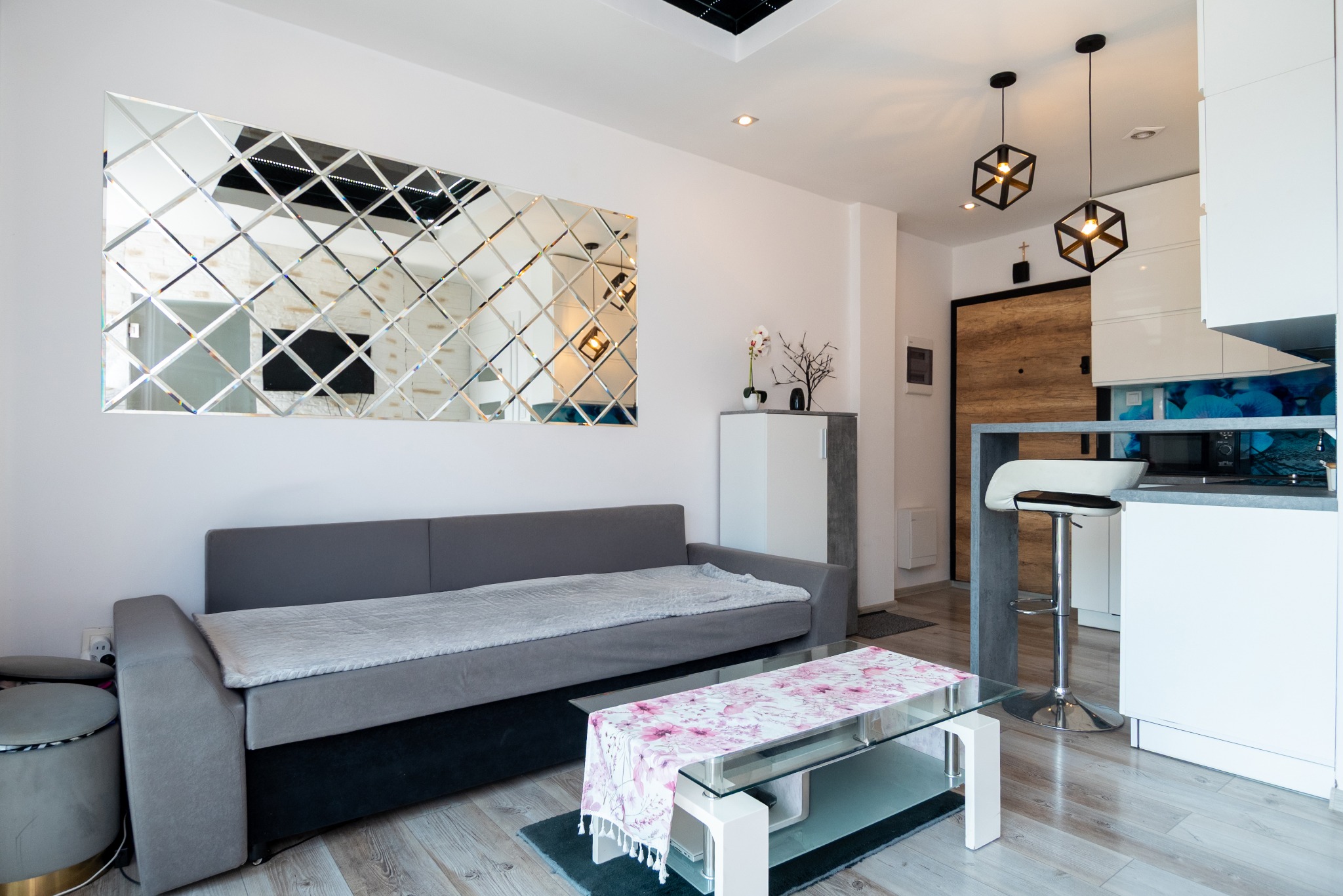 Nowoczesny salon z szarą sofą, lustrzaną ścianą i szklanym stolikiem kawowym. Oświetlenie stanowią geometryczne lampy wiszące. Widoczna kuchnia z barkiem i krzesłem.