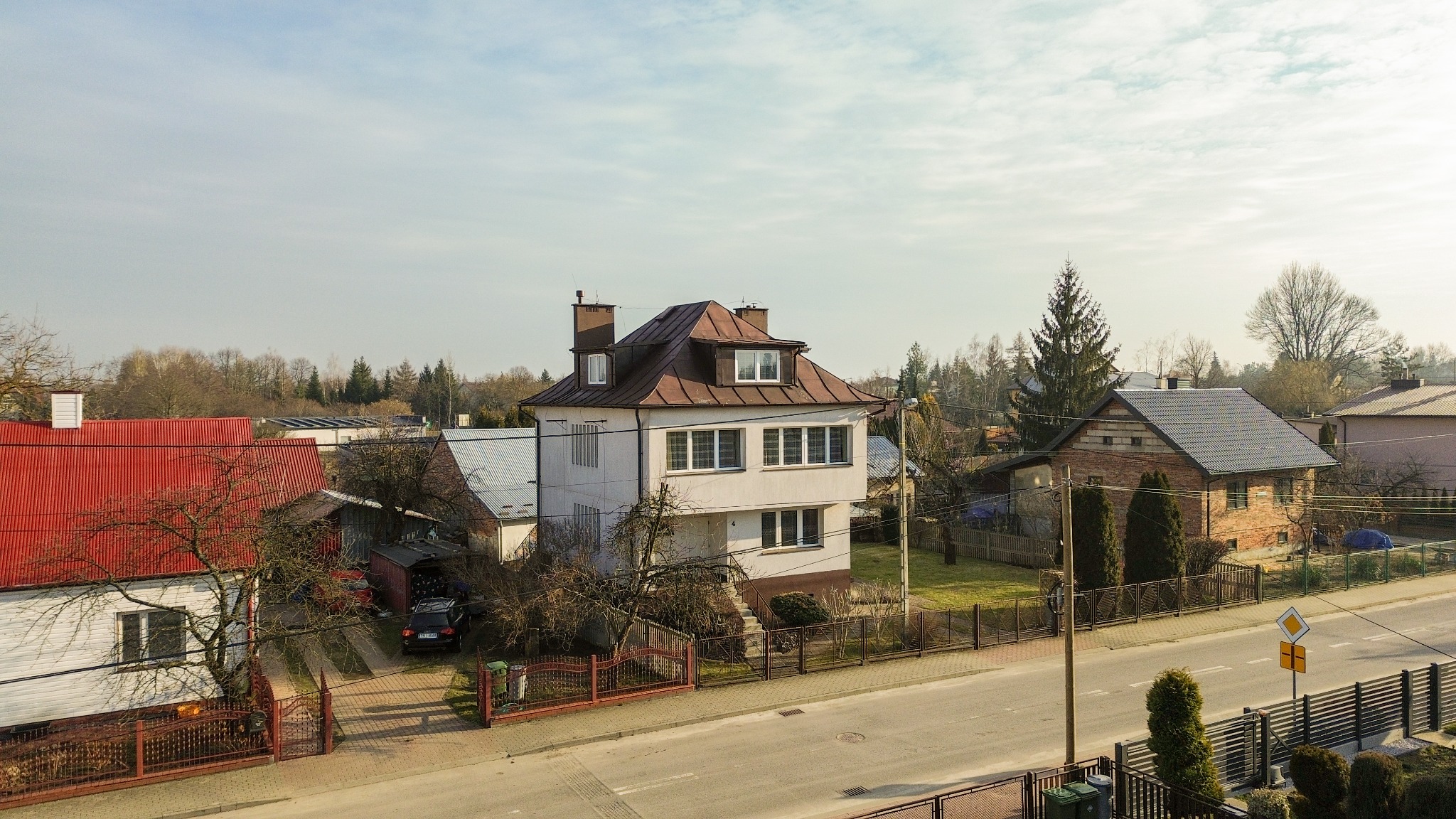 Dwupiętrowy dom z brązowym dachem, widziany z góry, w otoczeniu innych domów i drzew. Ulica przed posesją. Słoneczny dzień, jasne niebo. Widok z okna.