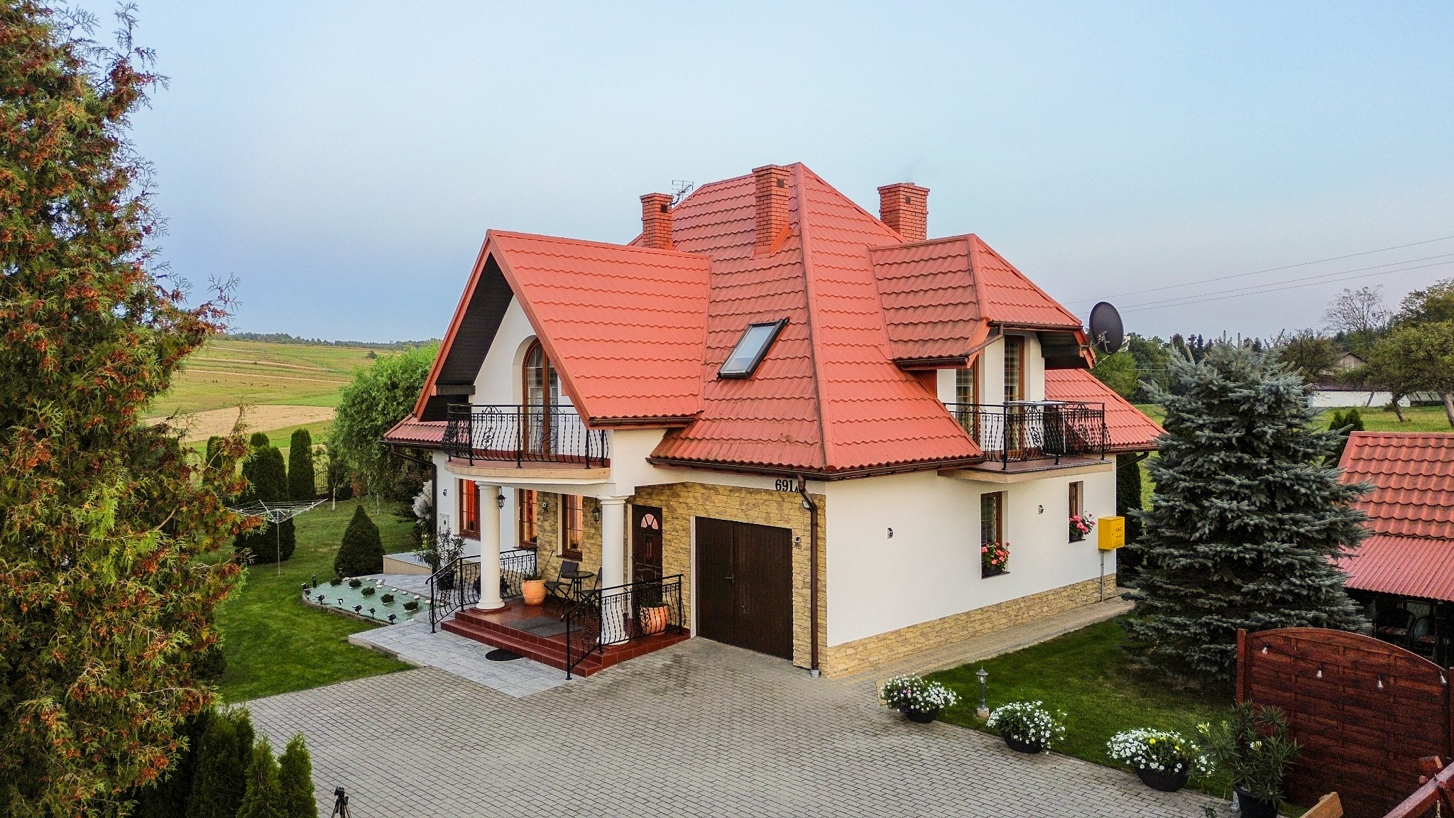 Dwupiętrowy dom z czerwonym dachem, balkonami z kutymi balustradami i garażem. Uwagę zwraca zadbany ogród z dekoracyjnymi krzewami i kostką brukową.