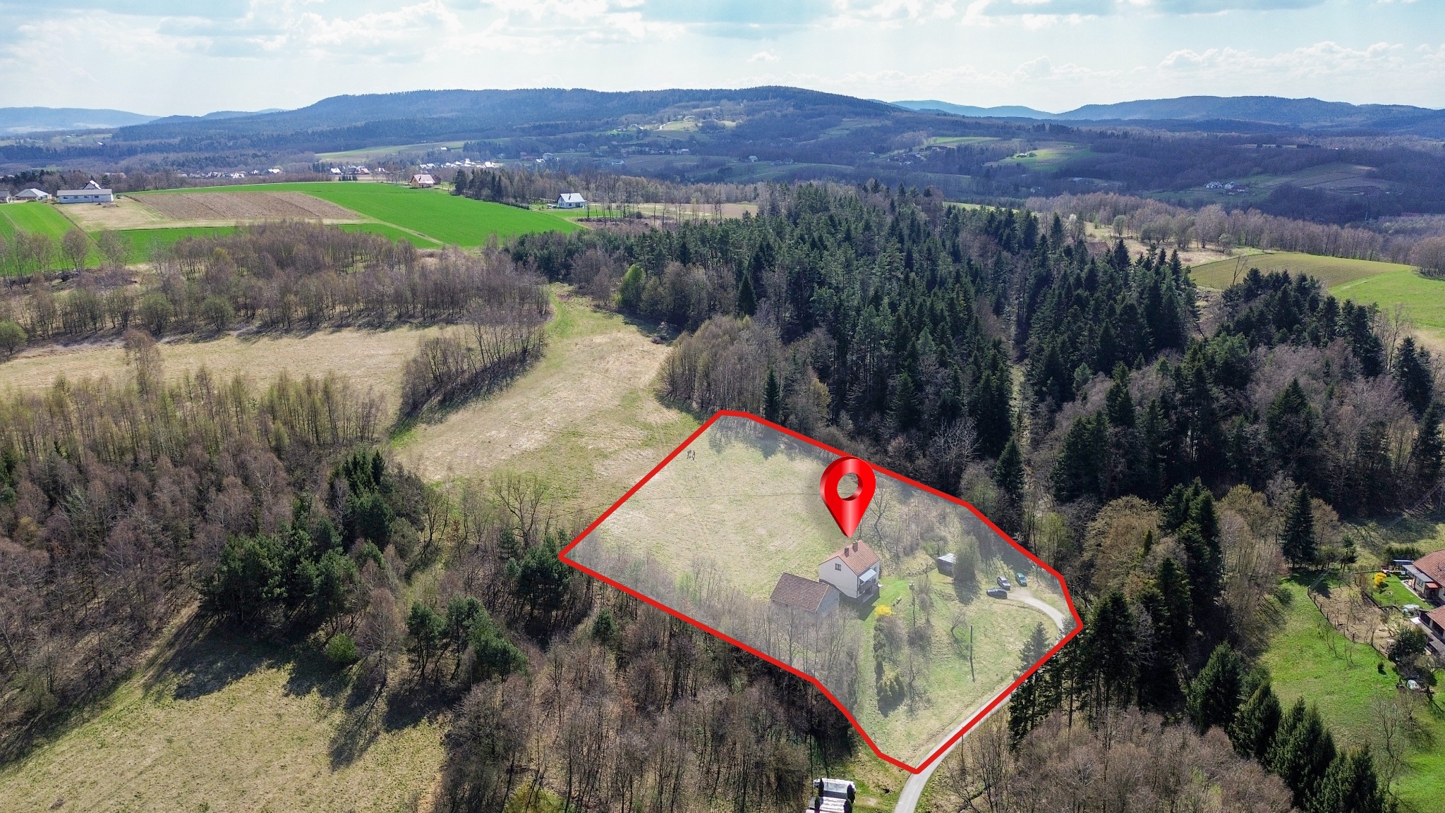 Widok z drona na dom z ogrodem, oznaczony czerwoną obwódką i pinezką lokalizacyjną, w otoczeniu lasów i pól. W tle pagórkowaty krajobraz.