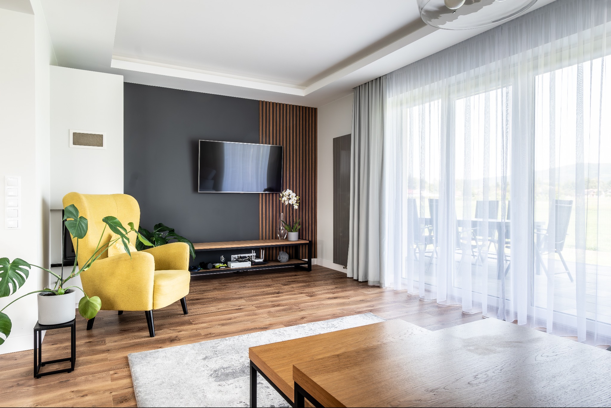 Jasne wnętrze salonu z żółtym fotelem, telewizorem na ścianie z drewnianymi panelami i dużym oknem z widokiem na zewnątrz, podkreślone naturalnym światłem.