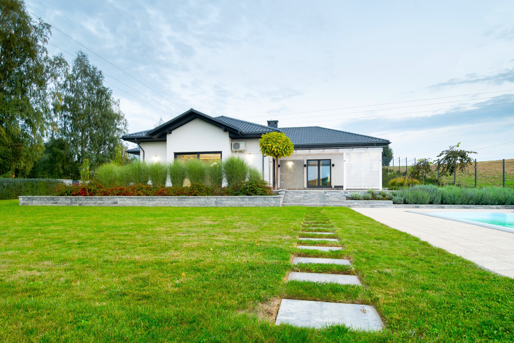 Nowoczesny dom jednorodzinny z zadbanym ogrodem, ścieżką z płyt i basenem w tle. Biała elewacja, ciemny dach, geometryczne nasadzenia.