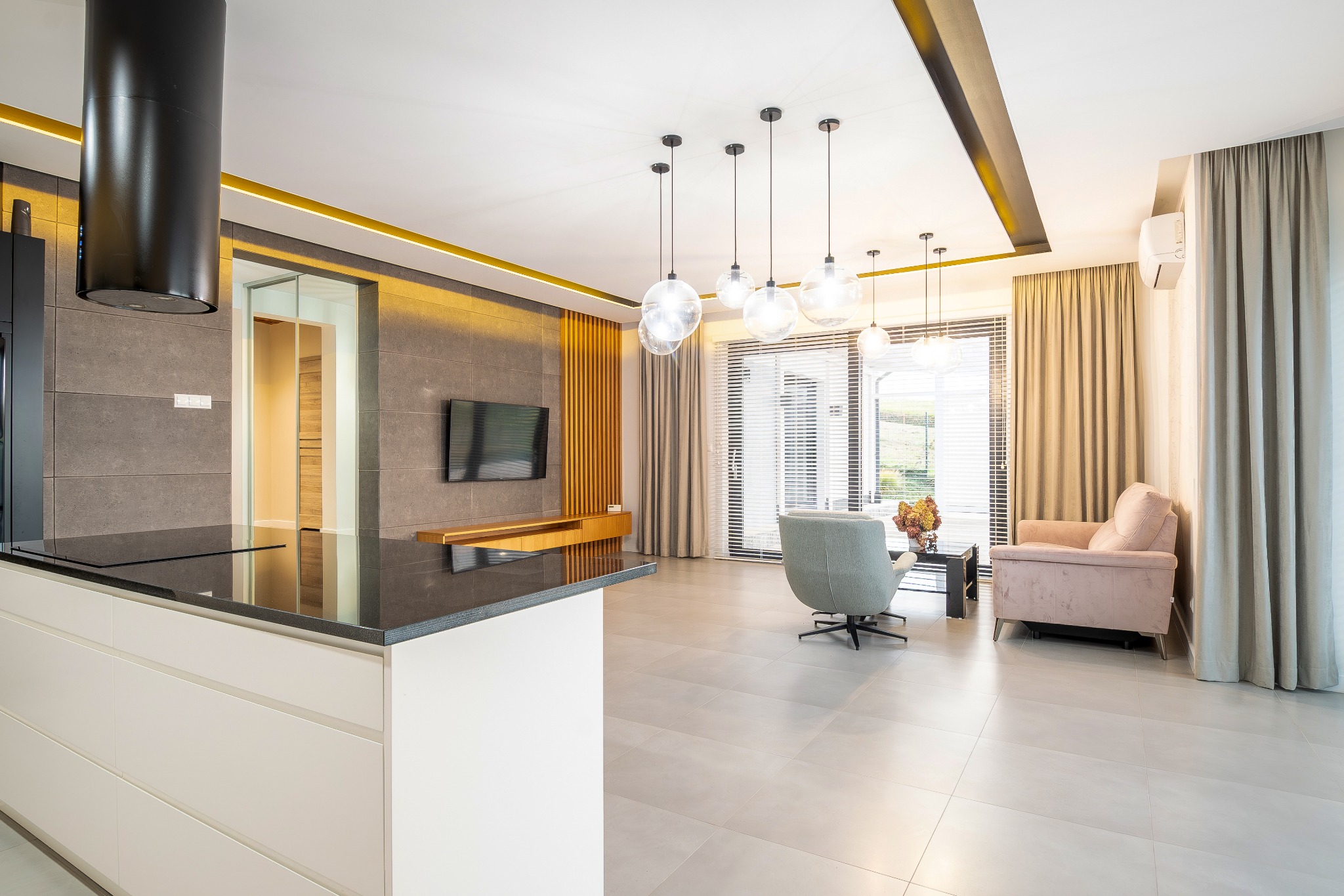 Nowoczesne wnętrze: otwarta kuchnia z wyspą, salon z szarym fotelem i różową sofą, duże okna z żaluzjami i wiszące lampy kuliste. Całość w jasnych barwach z akcentami złota.