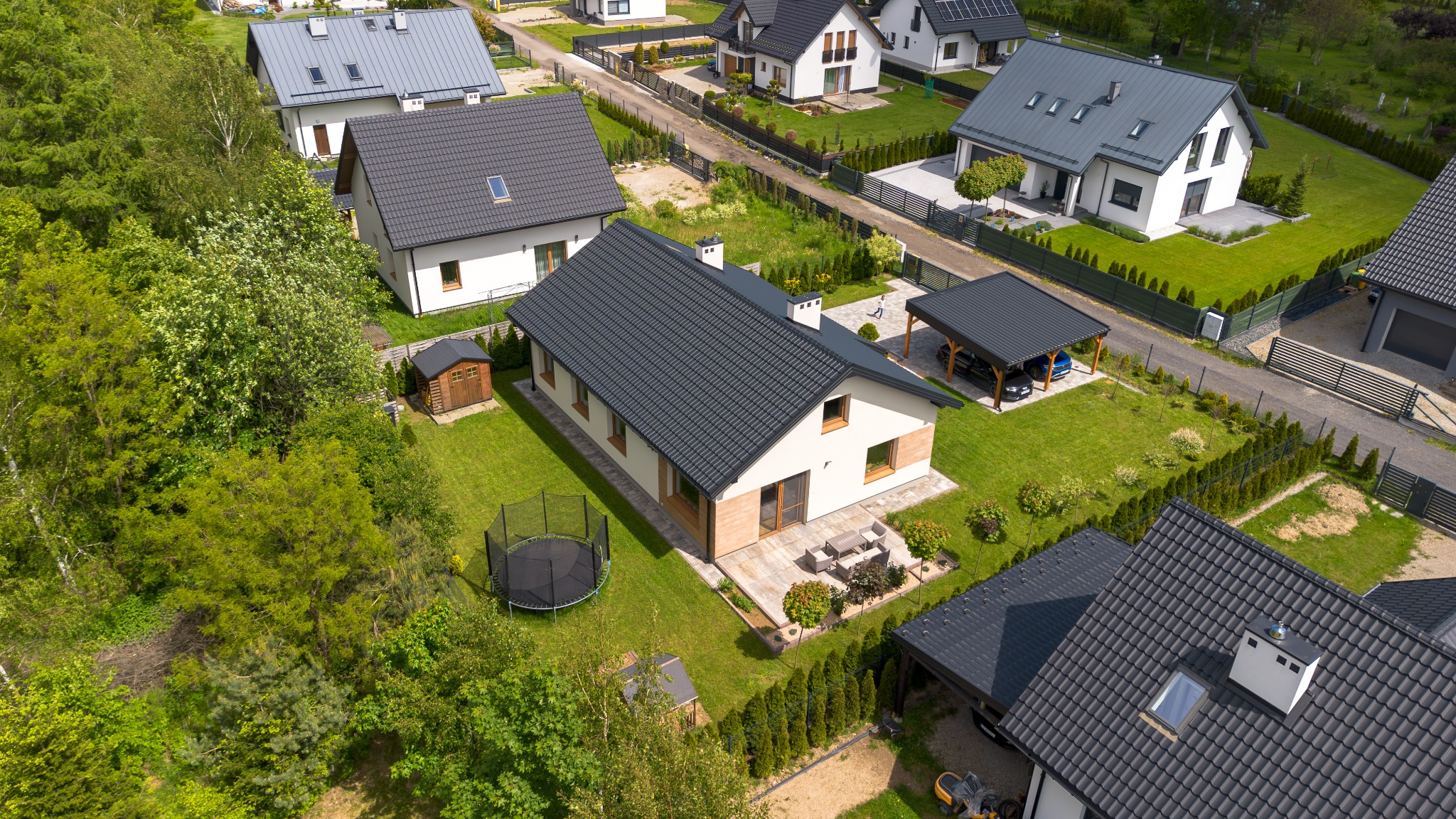 Widok z lotu ptaka na nowoczesny dom jednorodzinny z ciemnym dachem, zielonym trawnikiem, trampoliną i meblami ogrodowymi. W tle inne domy i drzewa.