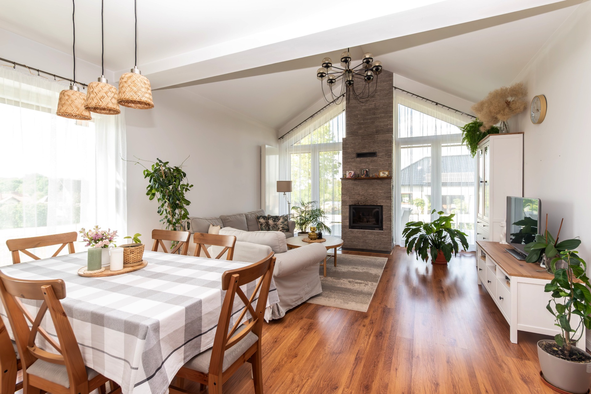 Jasne wnętrze domu z drewnianą podłogą, kominkiem z kamienia i stołem z krzesłami. W tle sofa i duże okna. Widoczne rośliny doniczkowe i lampy.