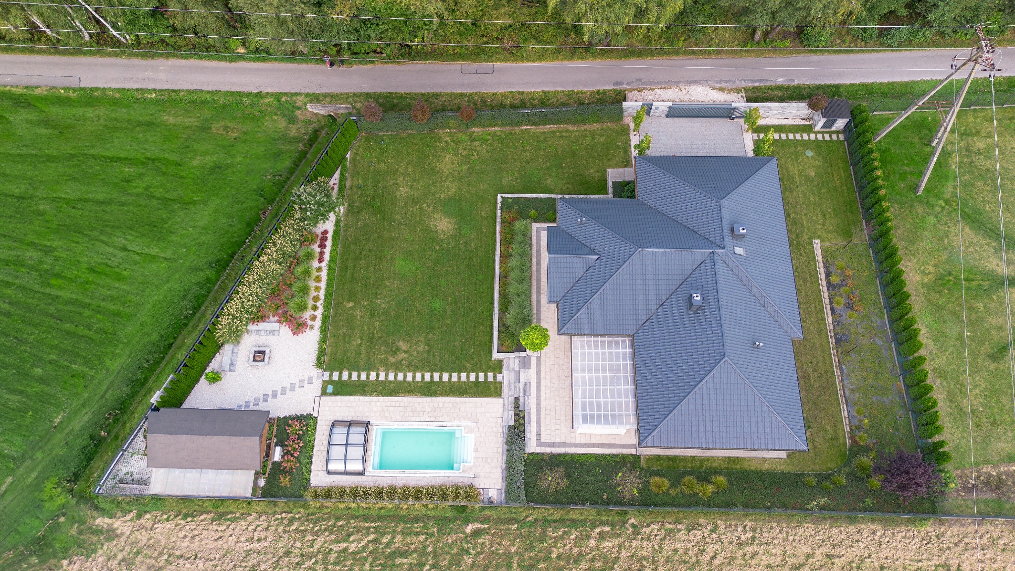 Widok z drona na duży dom z ogrodem, basenem i altaną. Regularne trawniki, geometryczne rabaty kwiatowe i ciemnoszary dach tworzą uporządkowaną całość.