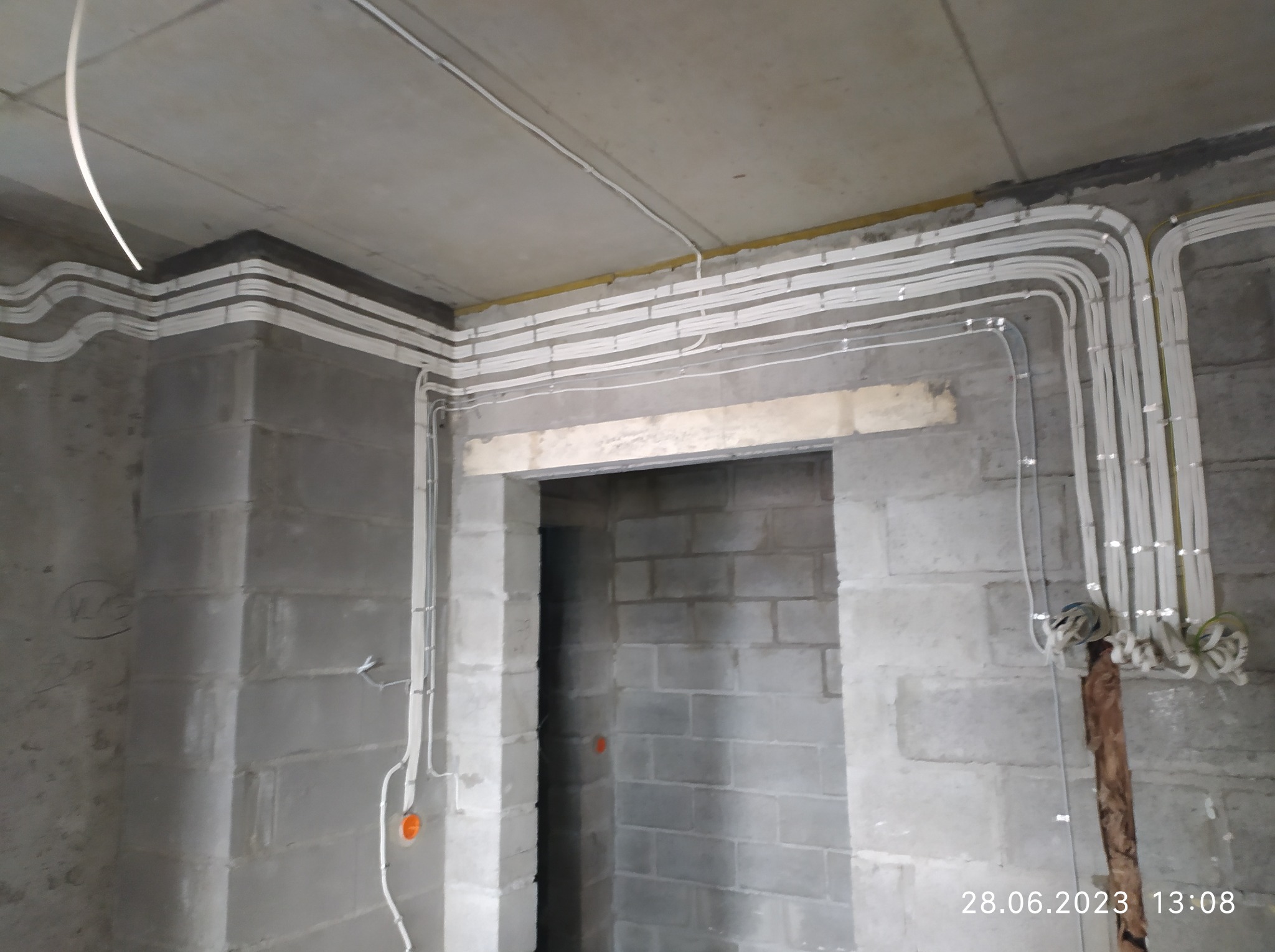 Instalacja elektryczna natynkowa w surowym wnętrzu: białe kable ułożone w równoległe wiązki pod sufitem i wzdłuż ścian z betonu komórkowego przed tynkowaniem, widoczne wejście do pomieszczenia.