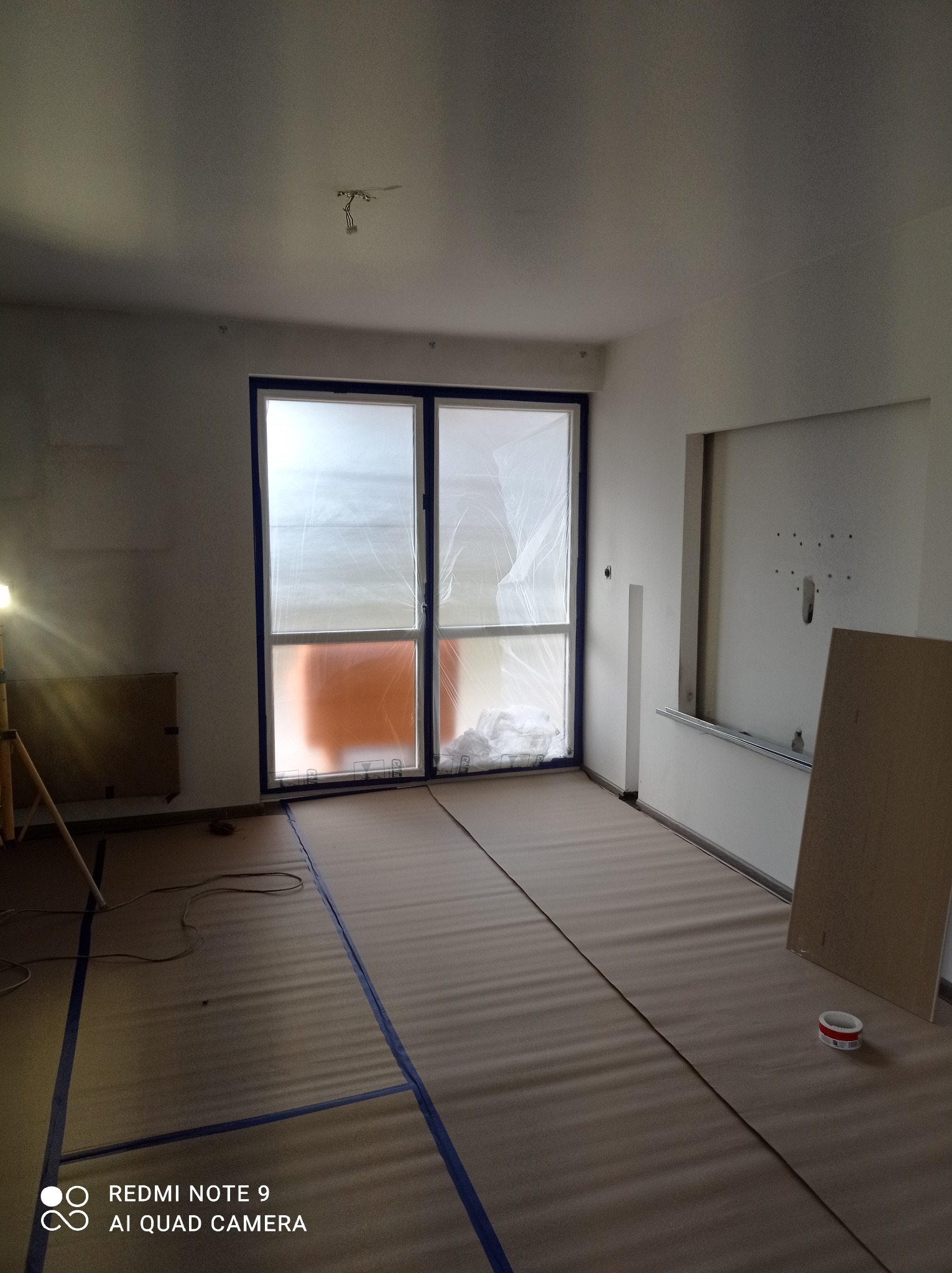 Przygotowanie do malowania pokoju: zabezpieczone okno folią, podłoga wyłożona kartonem i obklejona taśmą, widoczna puszka farby i lampa na statywie.