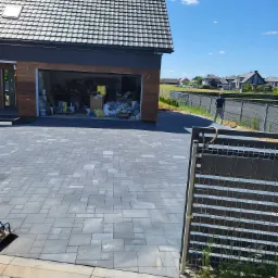 Ułożona szara kostka brukowa na podjeździe do garażu, widoczny fragment ogrodzenia i osoba z wężem ogrodowym w tle, brama garażowa otwarta z widocznym bałaganem w środku.