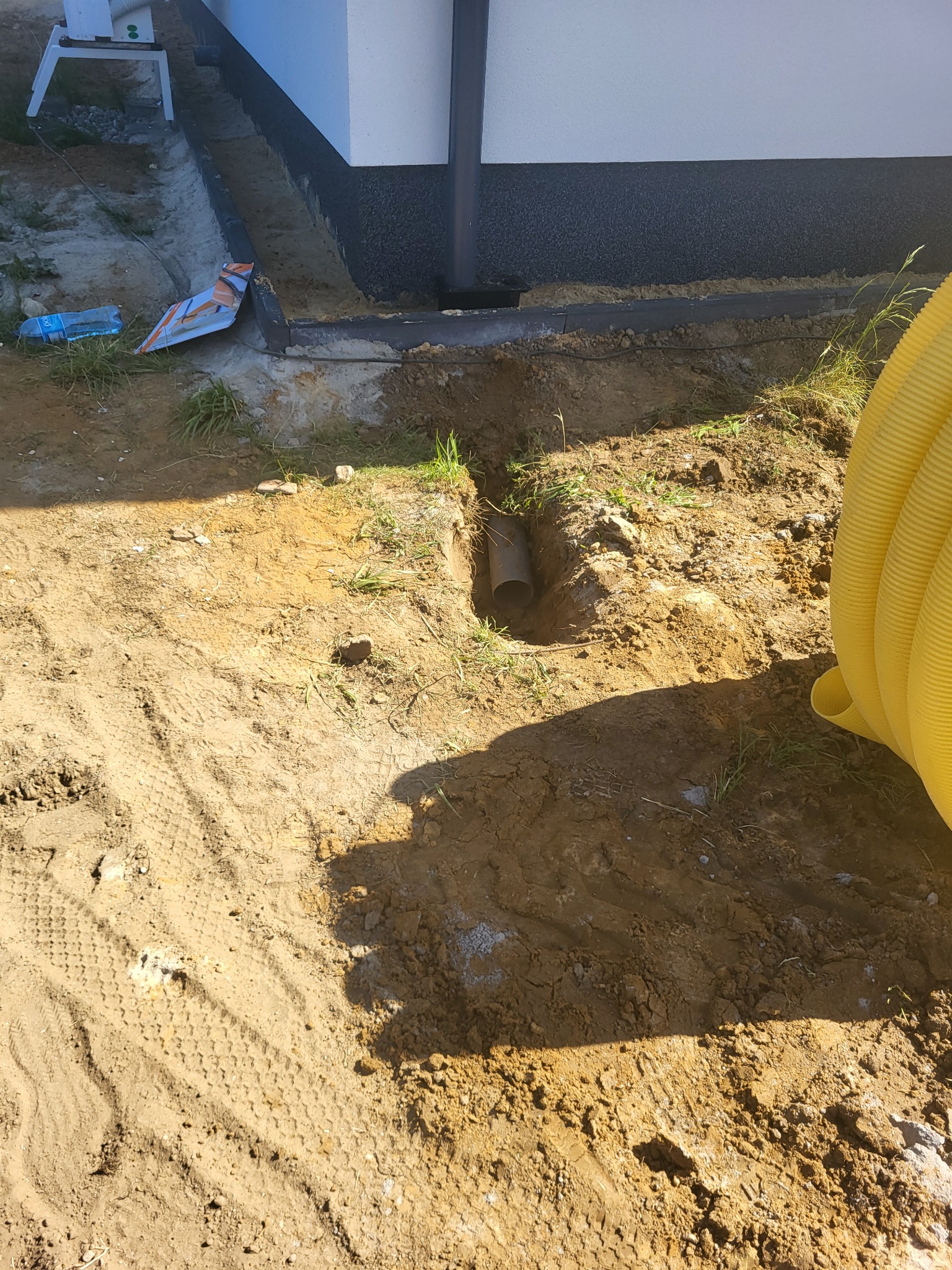 Wykop w ziemi z widoczną rurą odpływową, żółta rura karbowana w tle, fragment fundamentu budynku w górnej części kadru.