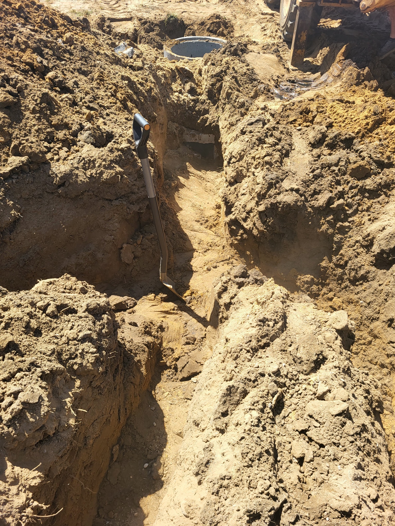 Wykopany rów w ziemi z betonowym kręgiem w tle, łopata wbita w dno, widoczny ślad opony ciężkiego sprzętu.