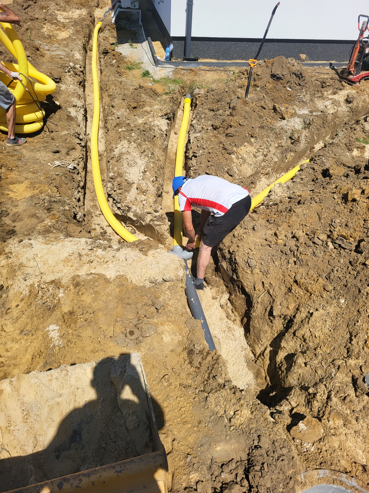 Instalacja żółtych rur osłonowych w wykopie pod fundamentami domu jednorodzinnego, widoczny pracownik w trakcie łączenia rur.