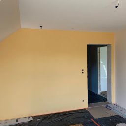 Proppo4k - Świeżo pomalowane na żółto ściany w pokoju na poddaszu, widoczne puszki po gniazdkach elektrycznych, podłoga zabezpieczona czarną folią, w tle widoczne przejście do innego pomieszczenia.