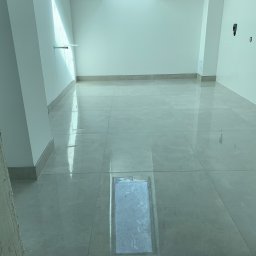 Proppo4k - Jasne, błyszczące płytki podłogowe w nowym pomieszczeniu z widoczną kratką wentylacyjną w podłodze, świeżo wykończone ściany i listwy przypodłogowe.