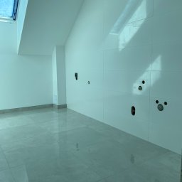 Proppo4k - Nowoczesne, jasne wnętrze z błyszczącymi, kwadratowymi płytkami na podłodze i ścianie, widoczne instalacje wodne i elektryczne w ścianie, okno dachowe w skośnym suficie.