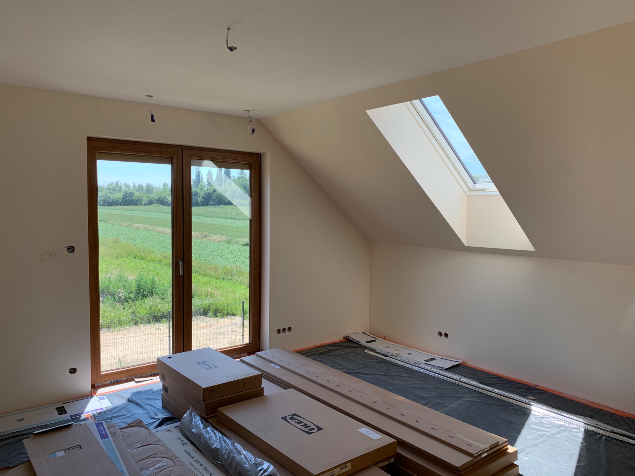 Poddasze w trakcie prac wykończeniowych: widok na okno dachowe, okno z widokiem na zieleń, przygotowane panele podłogowe w kartonach IKEA na czarnej folii zabezpieczającej.
