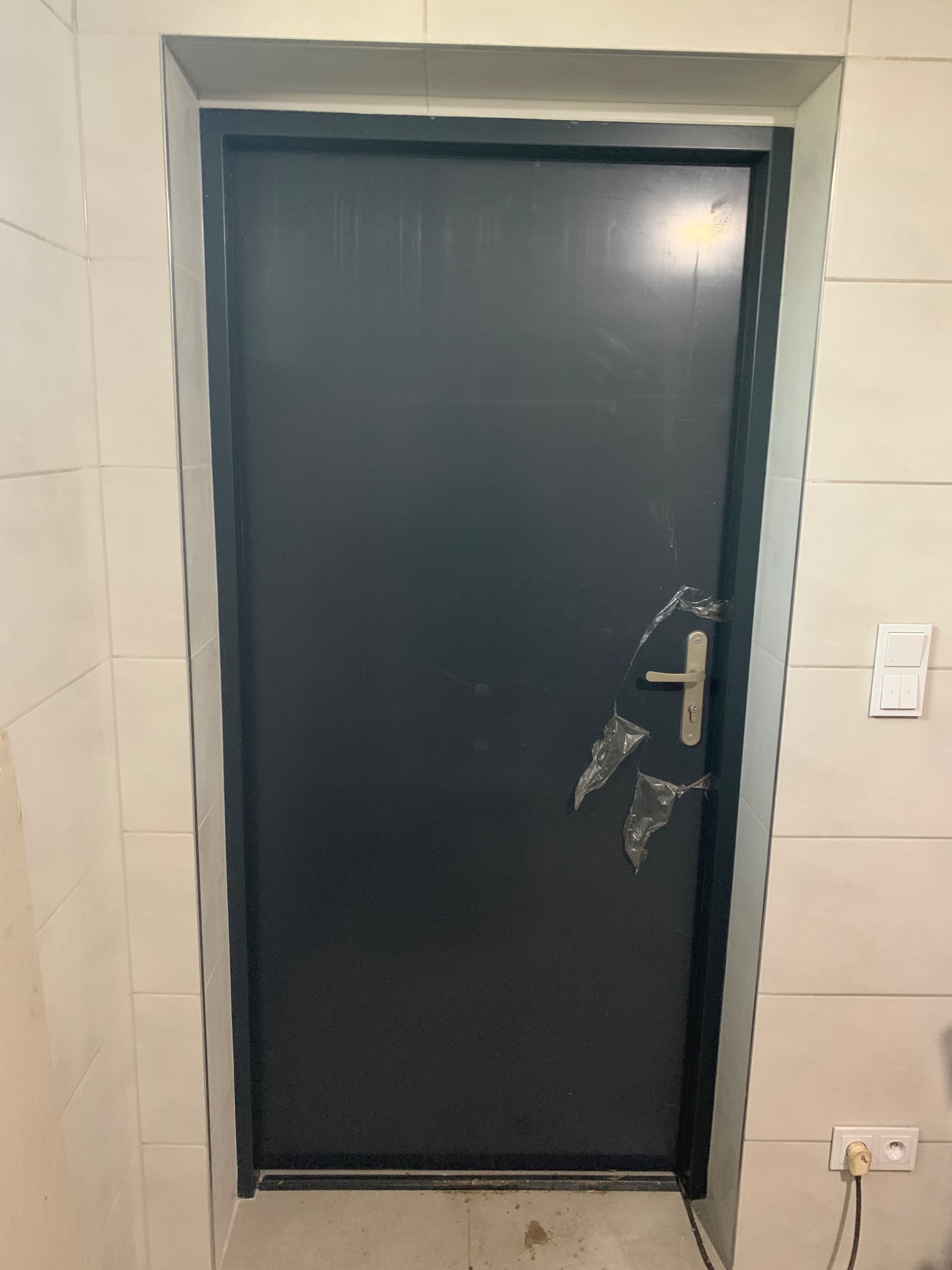 Nowe, czarne drzwi w łazience z białymi płytkami, zabezpieczone folią ochronną na klamce, widoczny kontakt i włącznik światła.