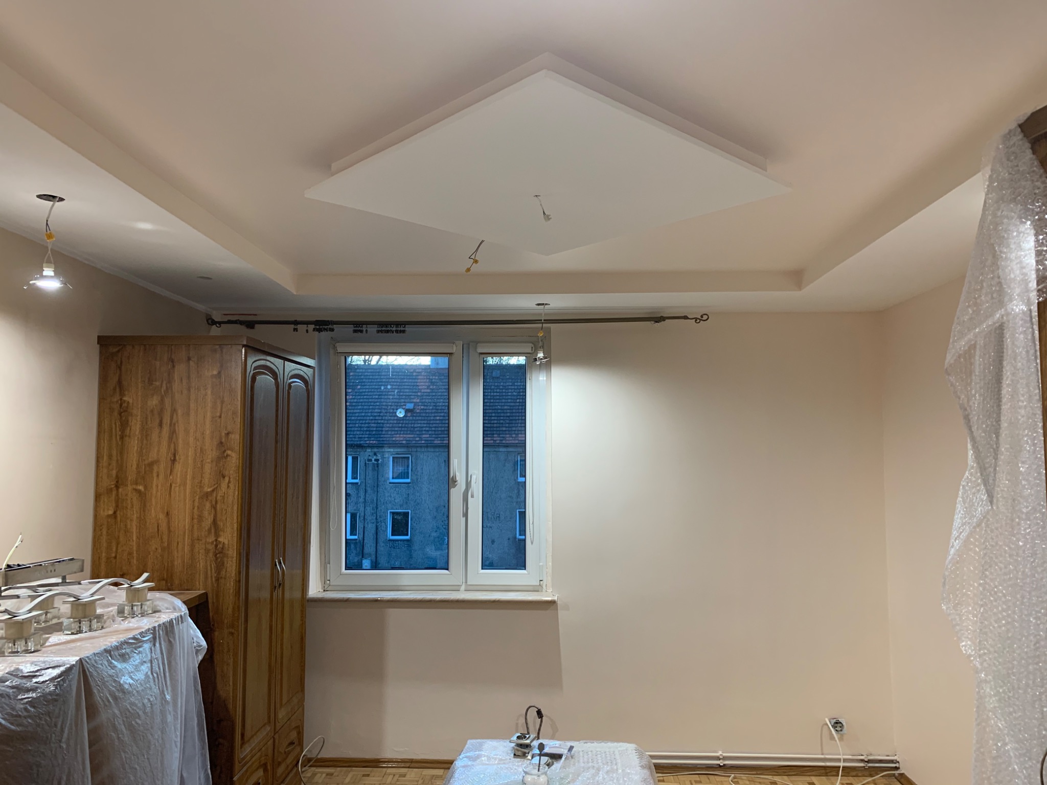 Pokój po odświeżeniu: pomalowane ściany w odcieniach beżu, nowy sufit podwieszany z centralnym kwadratowym panelem, drewniana szafa z widocznymi śladami użytkowania, okno z widokiem na budynek...