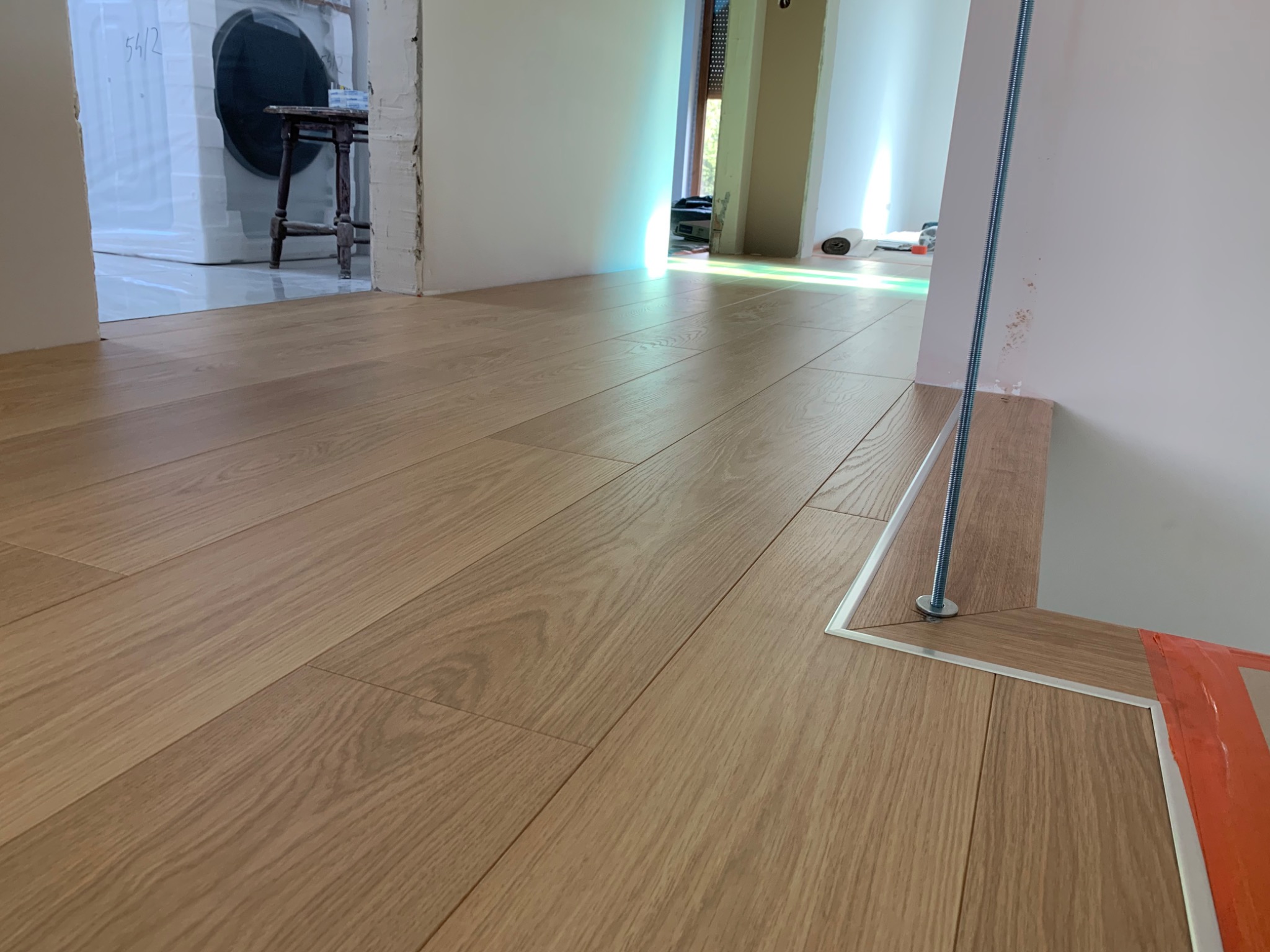 Jasny parkiet ułożony w nowym pomieszczeniu, widoczne listwy przypodłogowe i śruby mocujące, w tle sprzęt AGD owinięty folią ochronną.