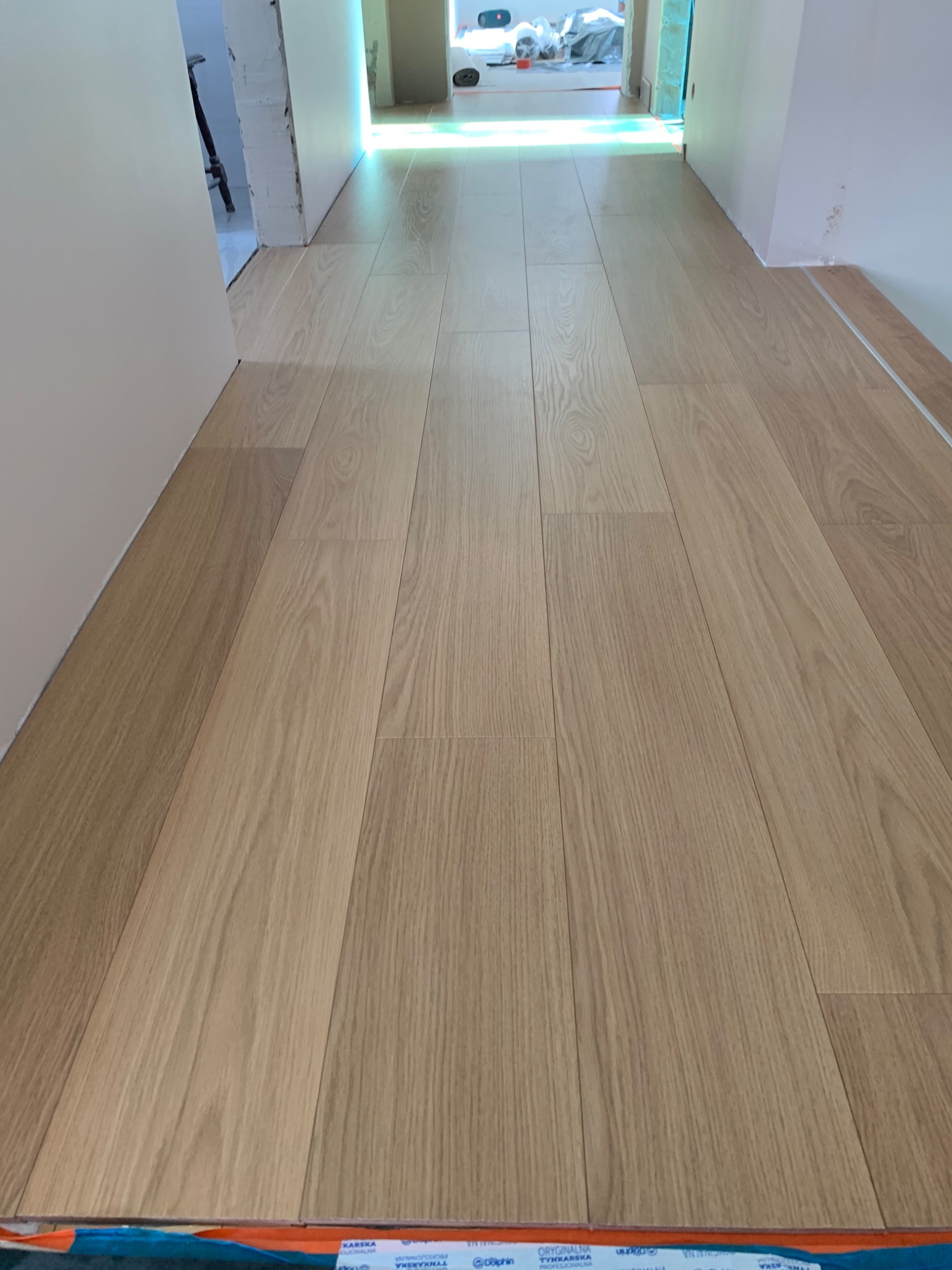 Jasne panele podłogowe ułożone w korytarzu, widoczne folie ochronne przy listwach przypodłogowych, w tle wejście do pomieszczenia z workami i sprzętem budowlanym.