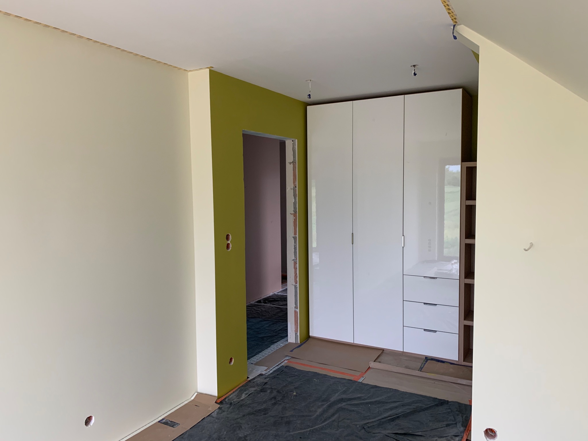 Wnętrze pokoju w trakcie prac wykończeniowych: pomalowane ściany w odcieniach beżu i oliwki, biała szafa z połyskiem, zabezpieczona podłoga, widoczne otwory na gniazdka elektryczne.