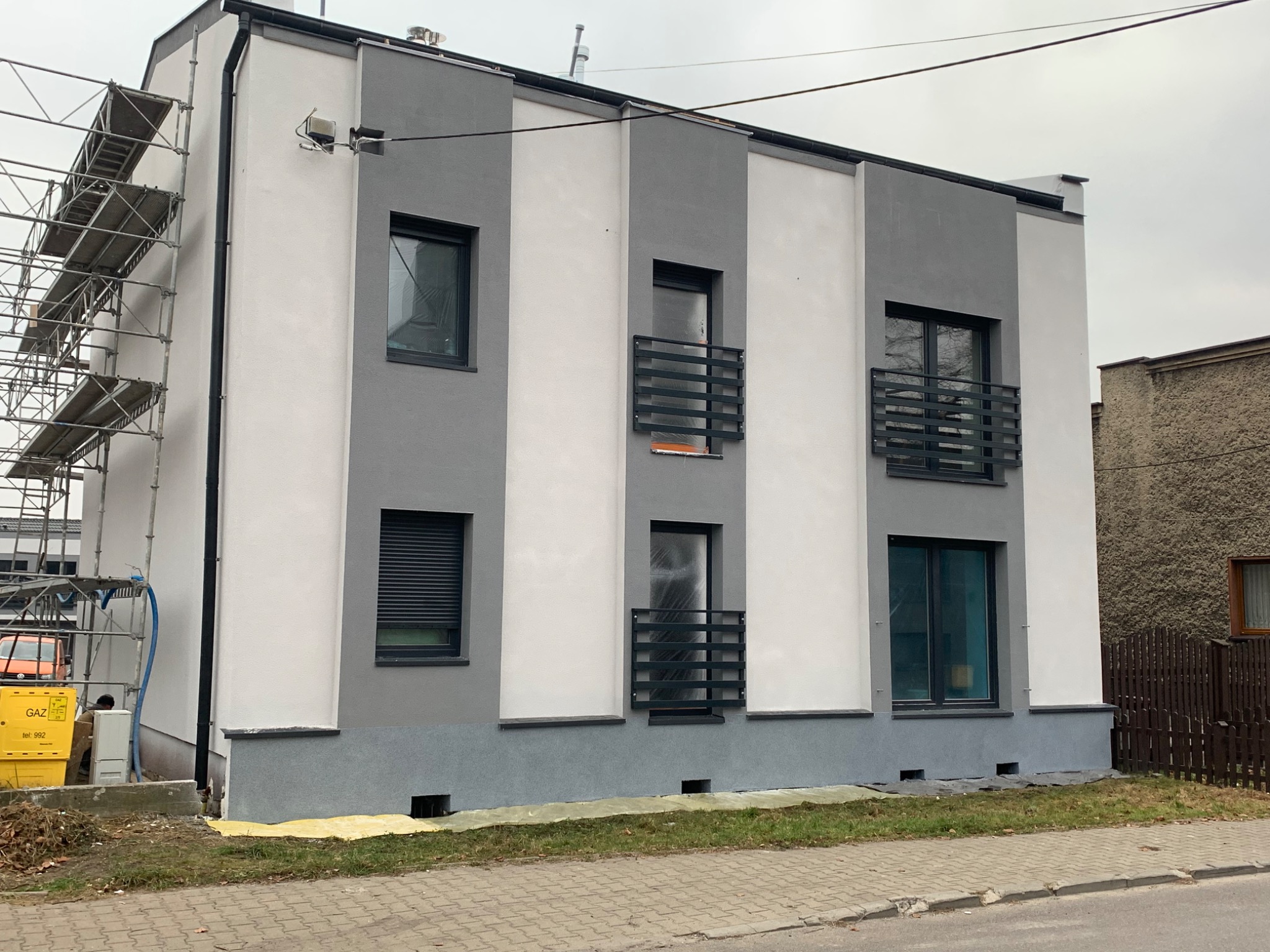 Dwukondygnacyjny budynek z szaro-białą elewacją w trakcie prac wykończeniowych, widoczne rusztowania i żółta folia ochronna na trawniku.