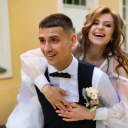 Uśmiechnięta panna młoda na barana u pana młodego, z bukiecikiem na jego klapie, na tle żółtej ściany z oknami.