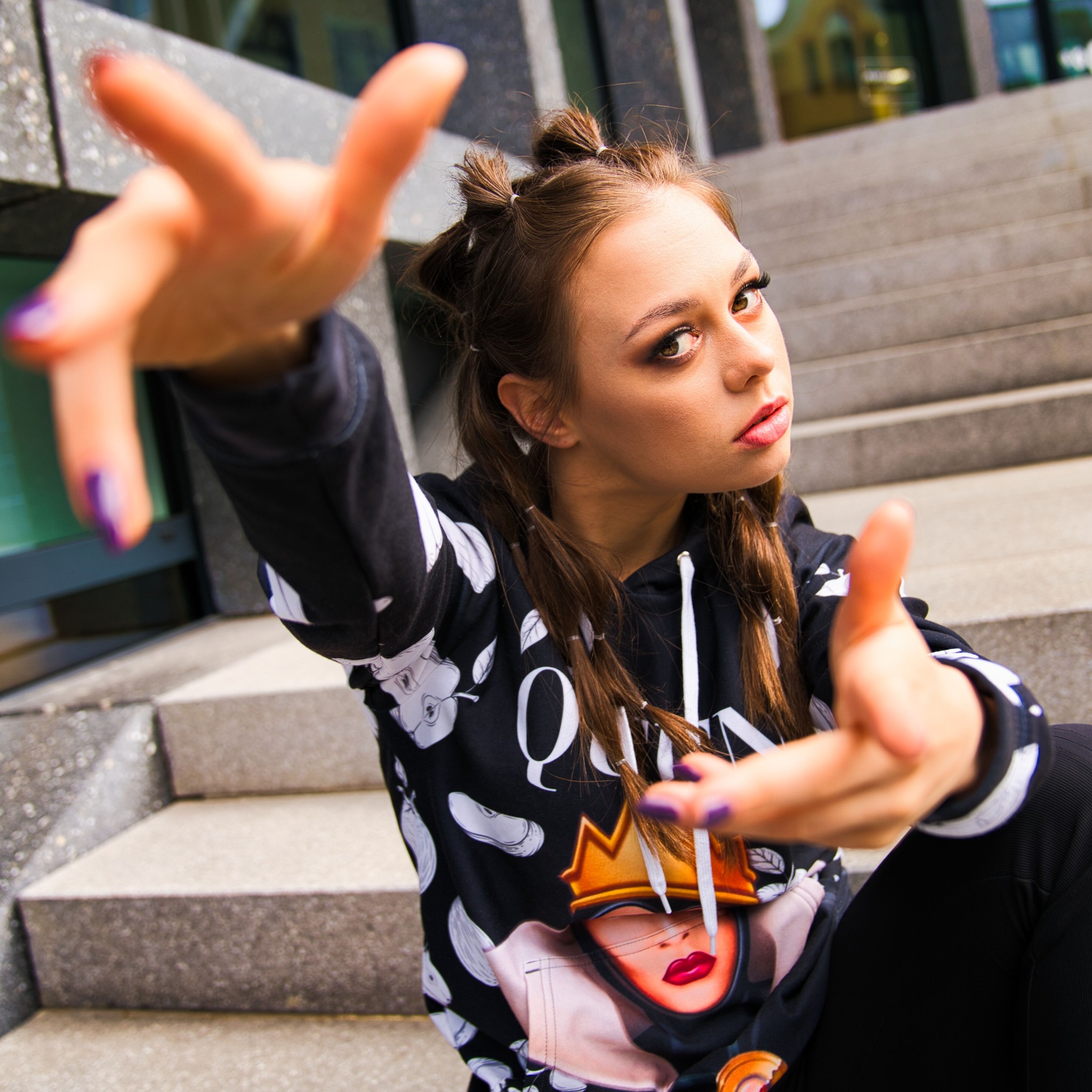 Młoda kobieta w czarnej bluzie z kapturem, siedząca na schodach, z wyciągniętymi dłońmi w kierunku obiektywu, prezentująca makijaż oczu i fioletowy manicure.