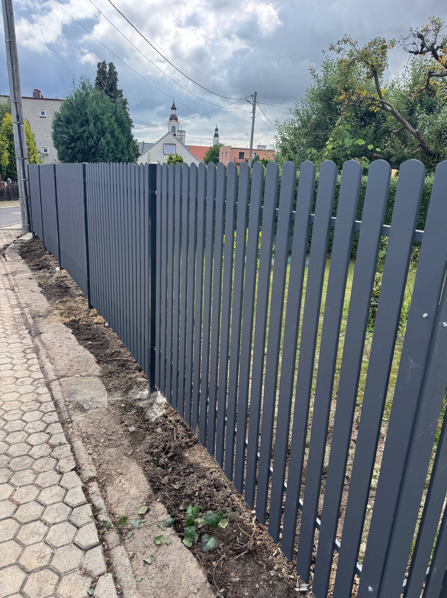 Nowoczesne, szare ogrodzenie panelowe z pionowych sztachet, świeżo zamontowane przy chodniku z kostki, z widocznym kościołem w tle. Minimalistyczny design.