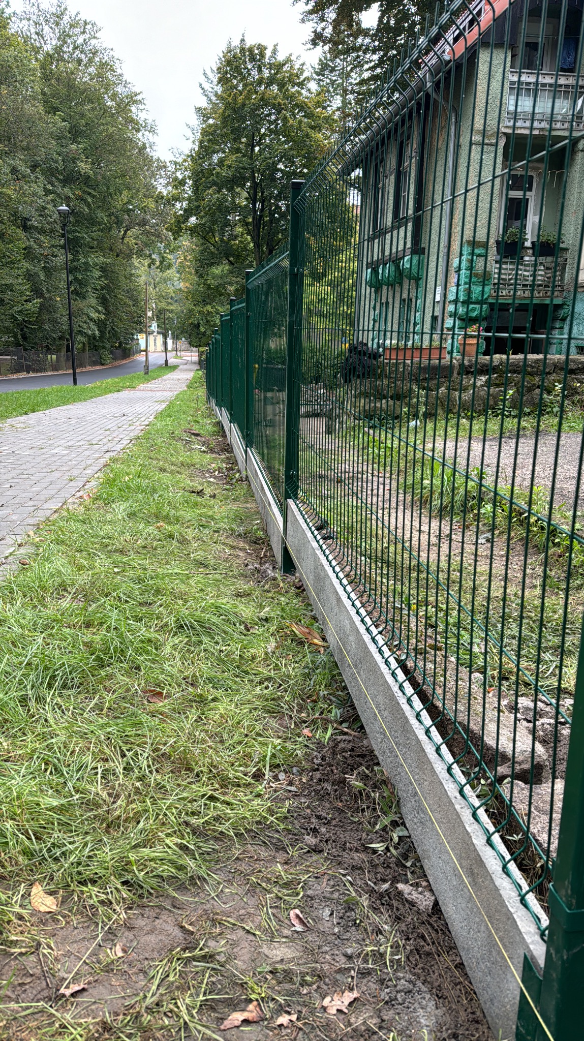 Zielone ogrodzenie panelowe z podmurówką betonową, rozciągające się wzdłuż trawnika i chodnika, z budynkiem w tle. Widoczna linka wyznaczająca linię ogrodzenia.