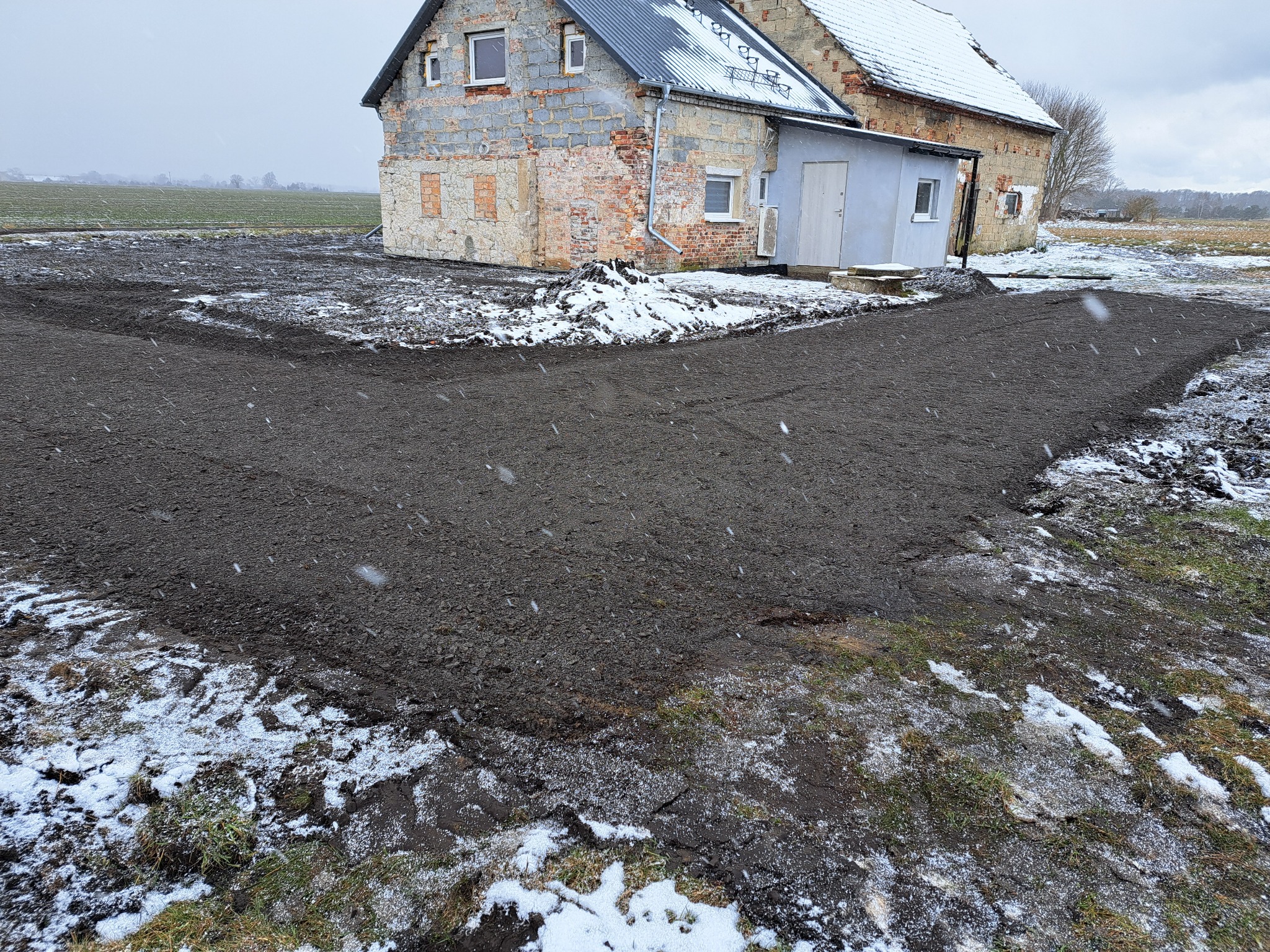 Przygotowany teren pod budowę lub nawierzchnię przy starym, ceglano-kamiennym budynku w zimowej scenerii, lekko ośnieżona ziemia, w tle pole i drzewa.