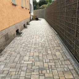 Ułożona kostka brukowa w geometryczny wzór, obok budynku z pomarańczową elewacją i gabionem z kamieni.