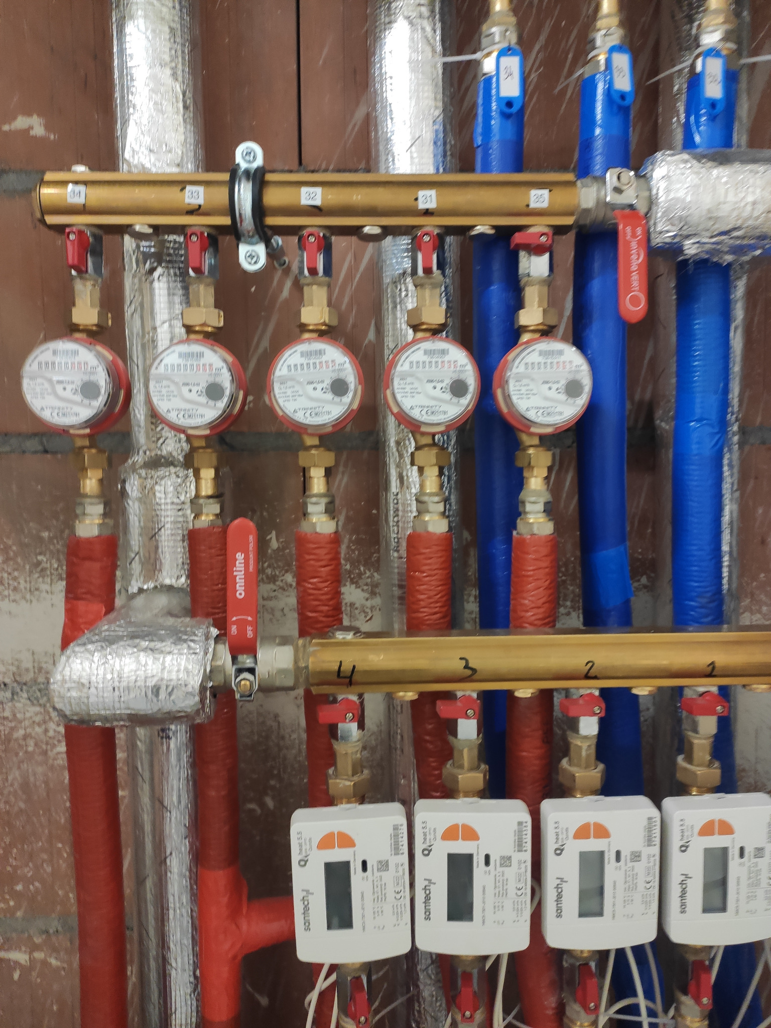 Rozdzielacz z licznikami ciepła Santech zamontowany na tle ceglanej ściany, widoczne rury w izolacji aluminiowej i niebieskie rury, z zaworami z czerwoną dźwignią.