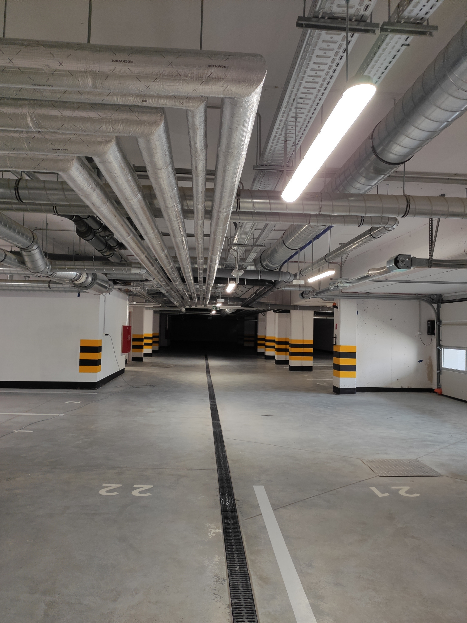 Instalacje wentylacyjne i rury pod sufitem w garażu podziemnym, widoczne oznaczenia stanowisk parkingowych i słupy z pasami ostrzegawczymi.