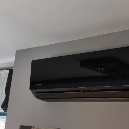 Czarny klimatyzator Rotenso Elis Series zamontowany na szarej ścianie w pomieszczeniu z roletą rzymską w oknie.