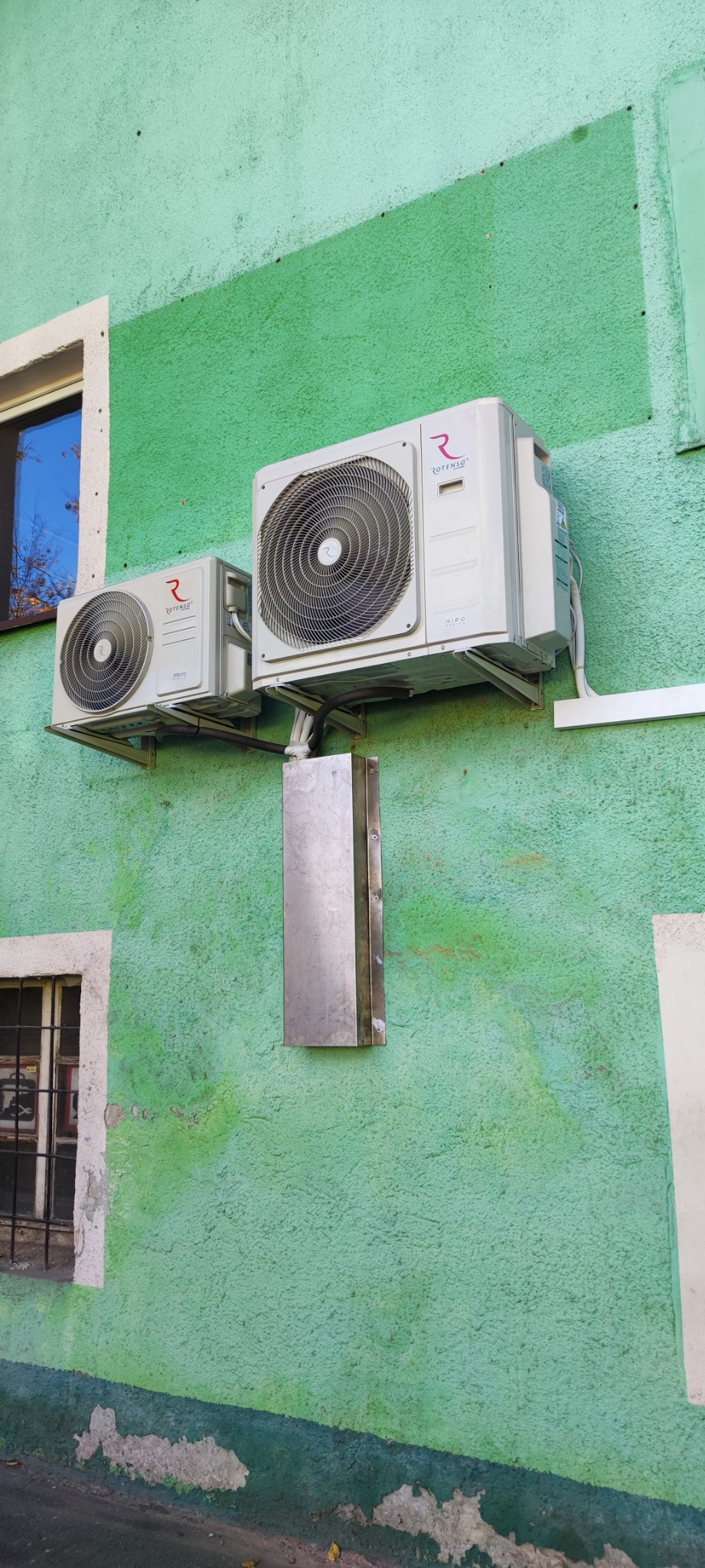 Dwa klimatyzatory marki Rotenso zamontowane na zielonej, teksturowanej ścianie budynku, widoczne okno z kratami w tle.