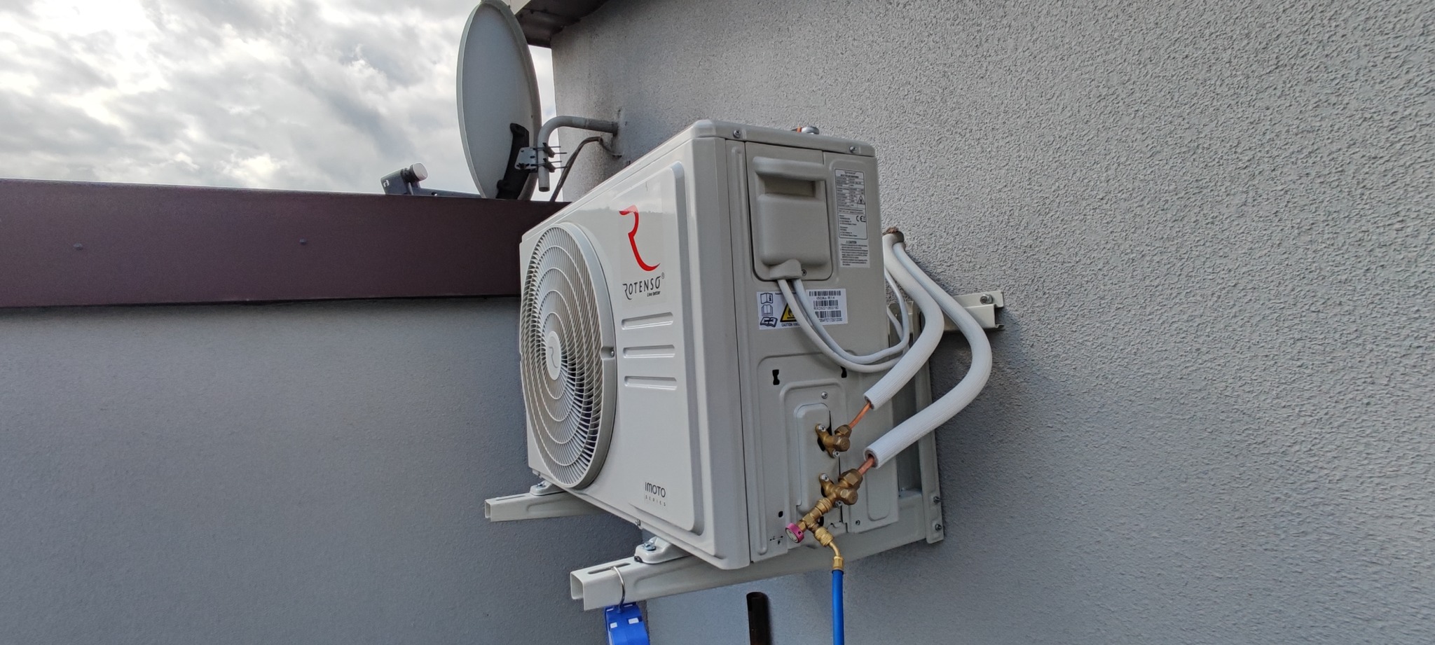 Zewnętrzna jednostka klimatyzacji Rotenso zamontowana na szarej elewacji budynku, widoczne rury połączeniowe i antena satelitarna na dachu w tle.
