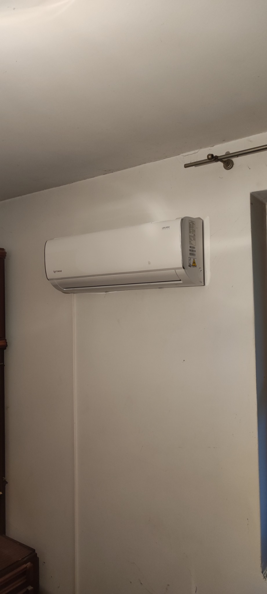 Biała jednostka wewnętrzna klimatyzatora Rotenso Likura zamontowana na ścianie w pomieszczeniu z widocznymi śladami użytkowania i niedoskonałościami na ścianie.