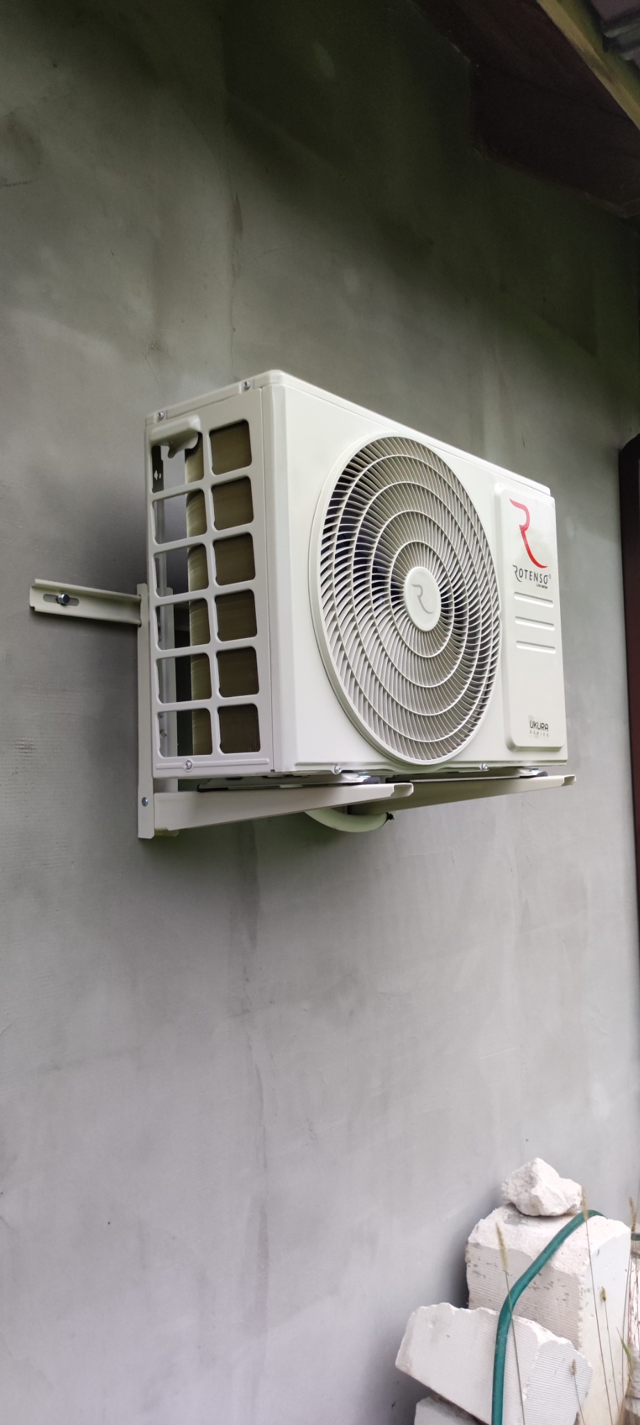 Zamontowana jednostka zewnętrzna klimatyzatora Rotenso Ukura na szarej, surowej ścianie zewnętrznej, z widocznymi fragmentami instalacji i stertą białych bloczków budowlanych u dołu.
