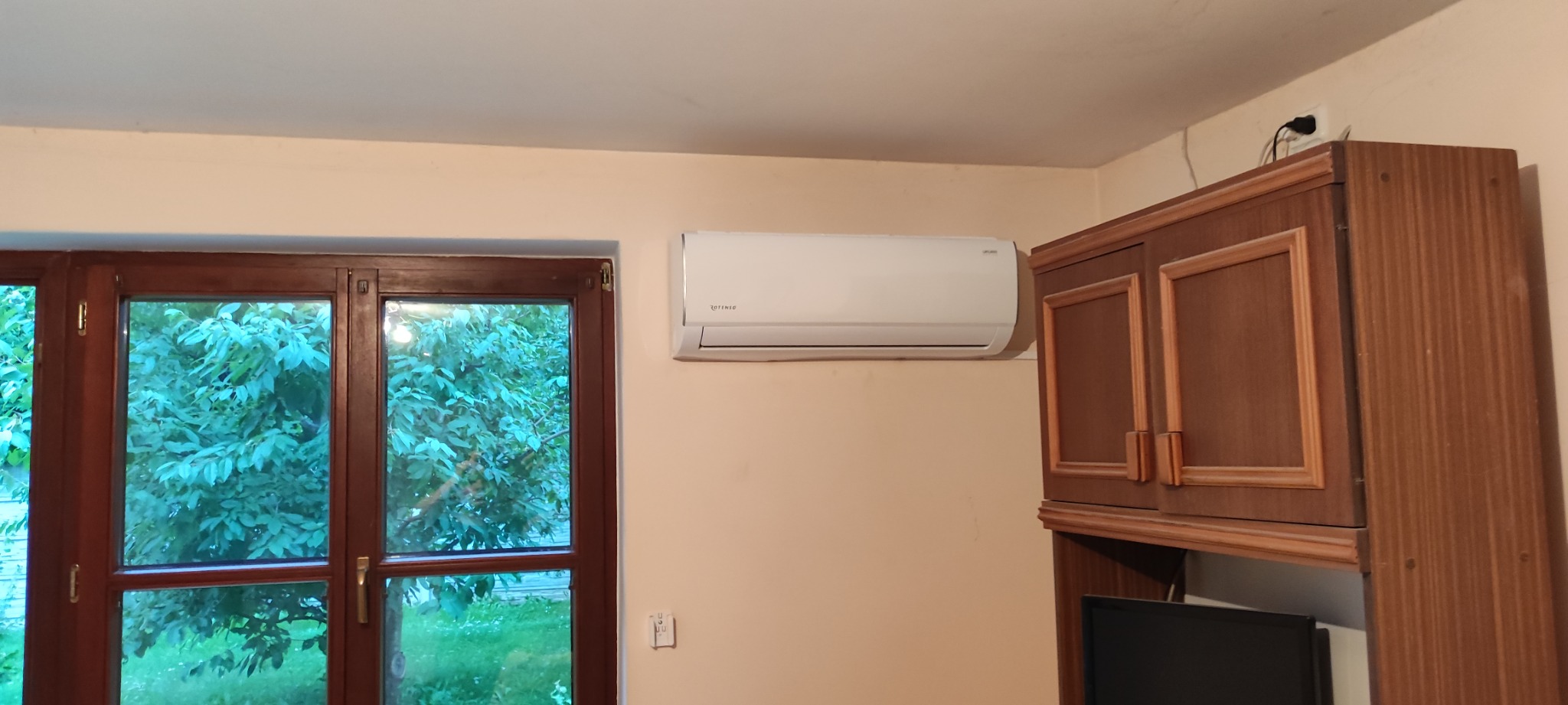 Biały klimatyzator marki Rotenso zamontowany na beżowej ścianie w pokoju, obok okna z drewnianą ramą i szafki z ciemnego drewna.