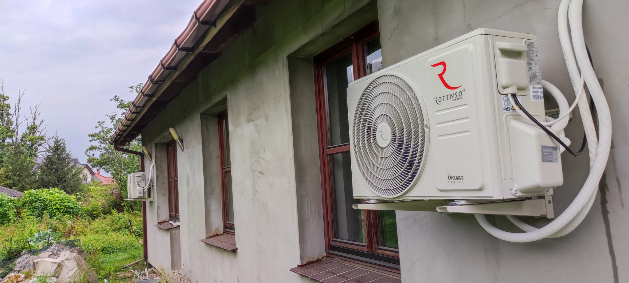 Zewnętrzna jednostka klimatyzacji Rotenso Ukura Series zamontowana na elewacji domu obok okna z brązowymi ramami; widoczne białe rury instalacyjne.