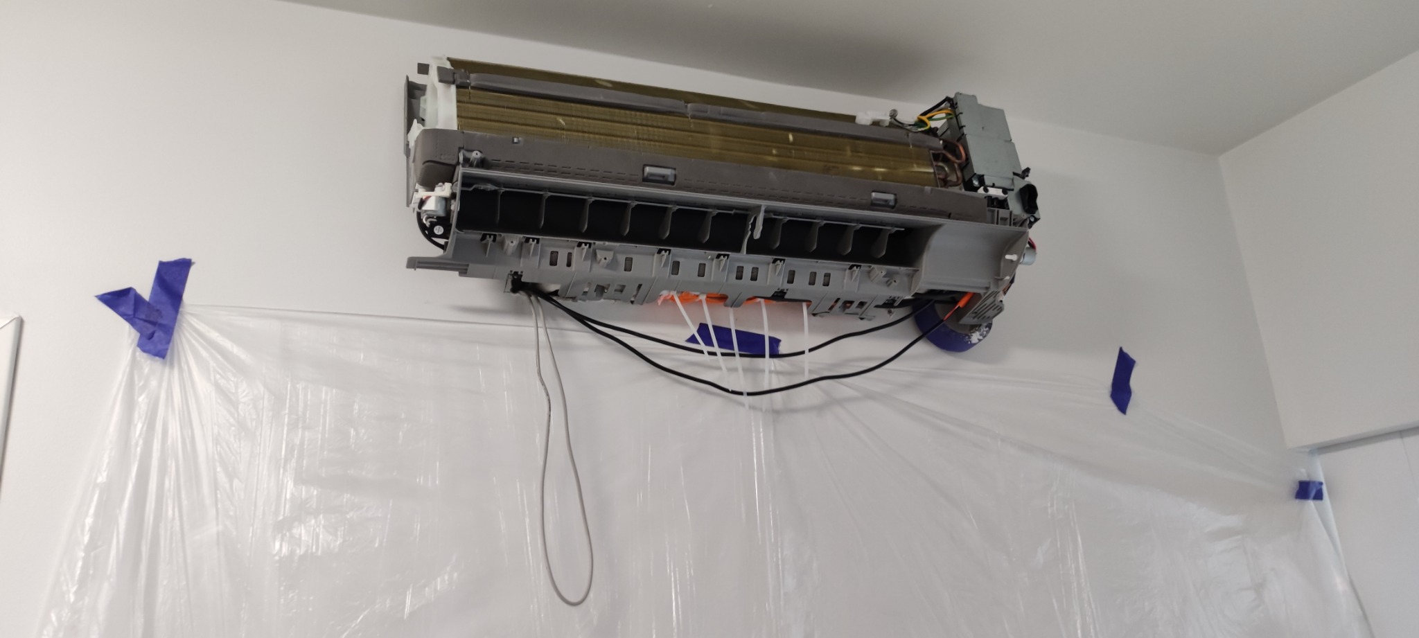 Rozłożona jednostka wewnętrzna klimatyzacji na ścianie, zabezpieczona folią ochronną przyklejoną niebieską taśmą malarską, widoczne przewody i elementy konstrukcyjne urządzenia.