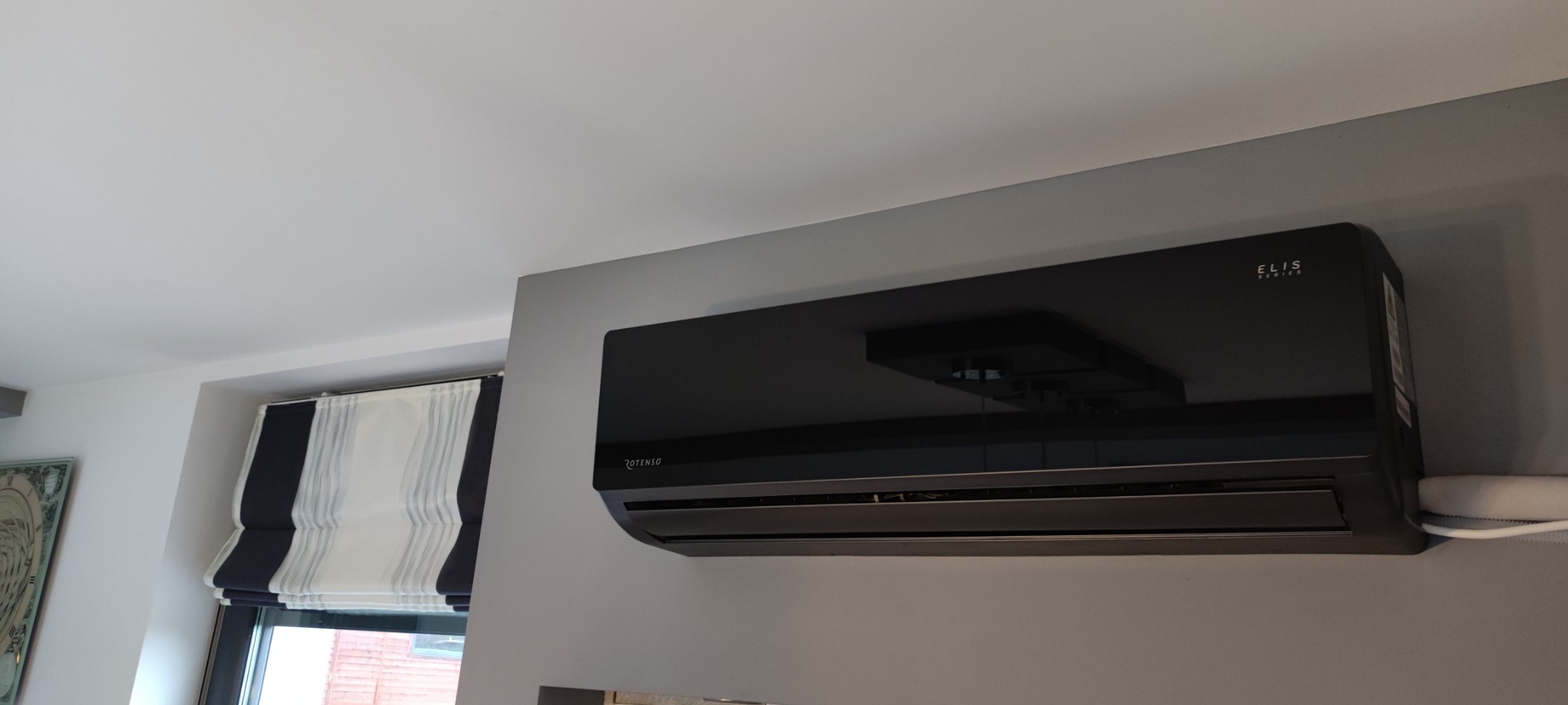 Czarny klimatyzator Rotenso Elis Series zamontowany na szarej ścianie w pomieszczeniu z roletą rzymską w oknie.