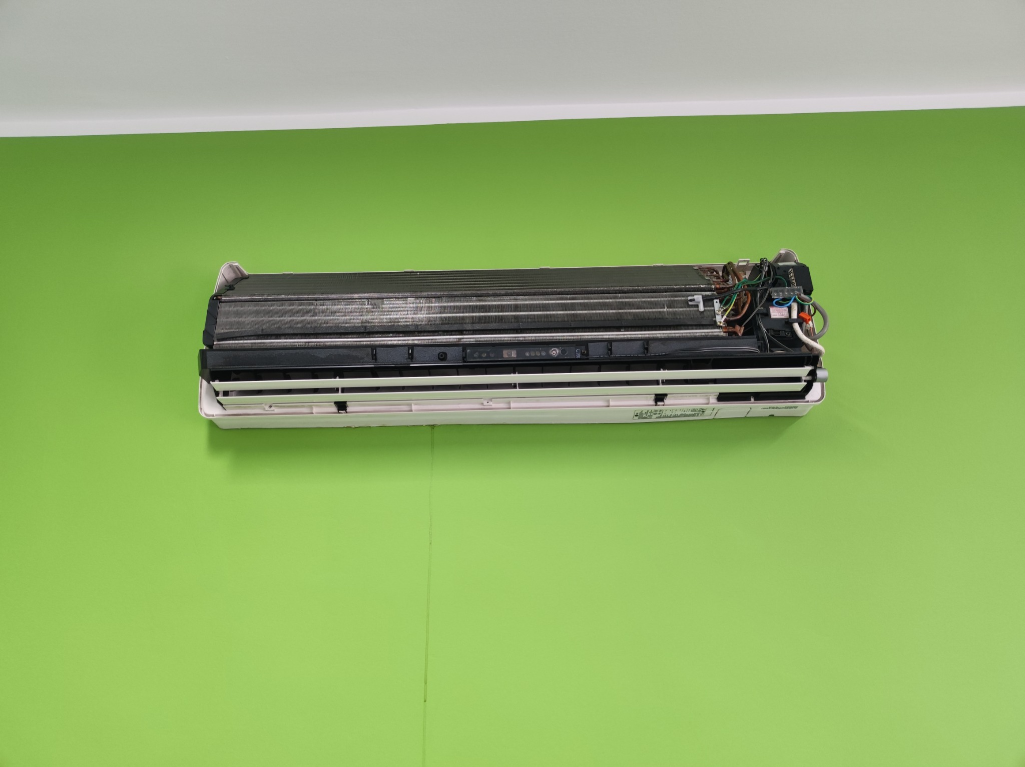 Otwarty klimatyzator ścienny z widocznymi elementami wewnętrznymi, zamontowany na zielonej ścianie.