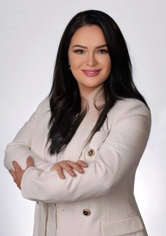 Portret uśmiechniętej kobiety w białym żakiecie z rękami skrzyżowanymi na tle jasnoszarego tła.