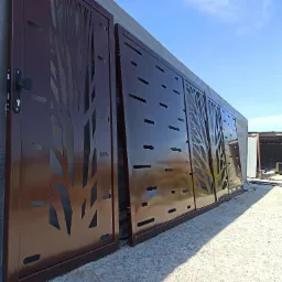 Brązowa brama wjazdowa z laserowo wycinanym wzorem drzewa, zamontowana na posesji z widocznym budynkiem gospodarczym w tle.