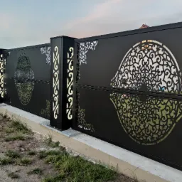 Nowoczesne, czarne ogrodzenie panelowe z ażurowymi, dekoracyjnymi wycięciami w kształcie stylizowanych kwiatów i ornamentów, podświetlone kolumny, widok z poziomu gruntu.