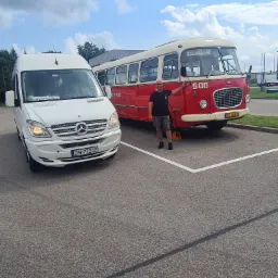 Biały Mercedes-Benz Sprinter z napisem 'Turystyczny' za szybą, obok czerwono-białego zabytkowego autobusu '500' z mężczyzną opierającym się o niego, zaparkowane na parkingu z wyznaczonymi miejscami.